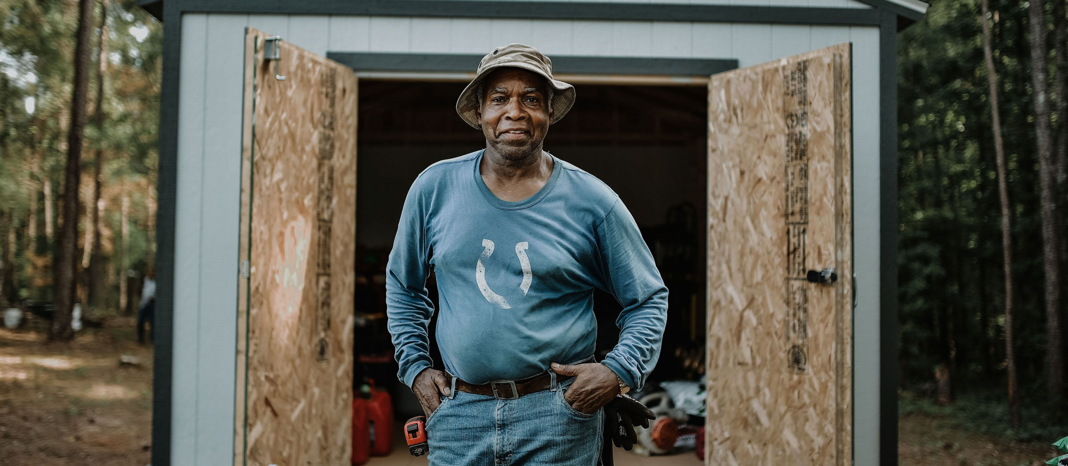 Man with his hands on his hips smiling at camera standing outside his shed. 